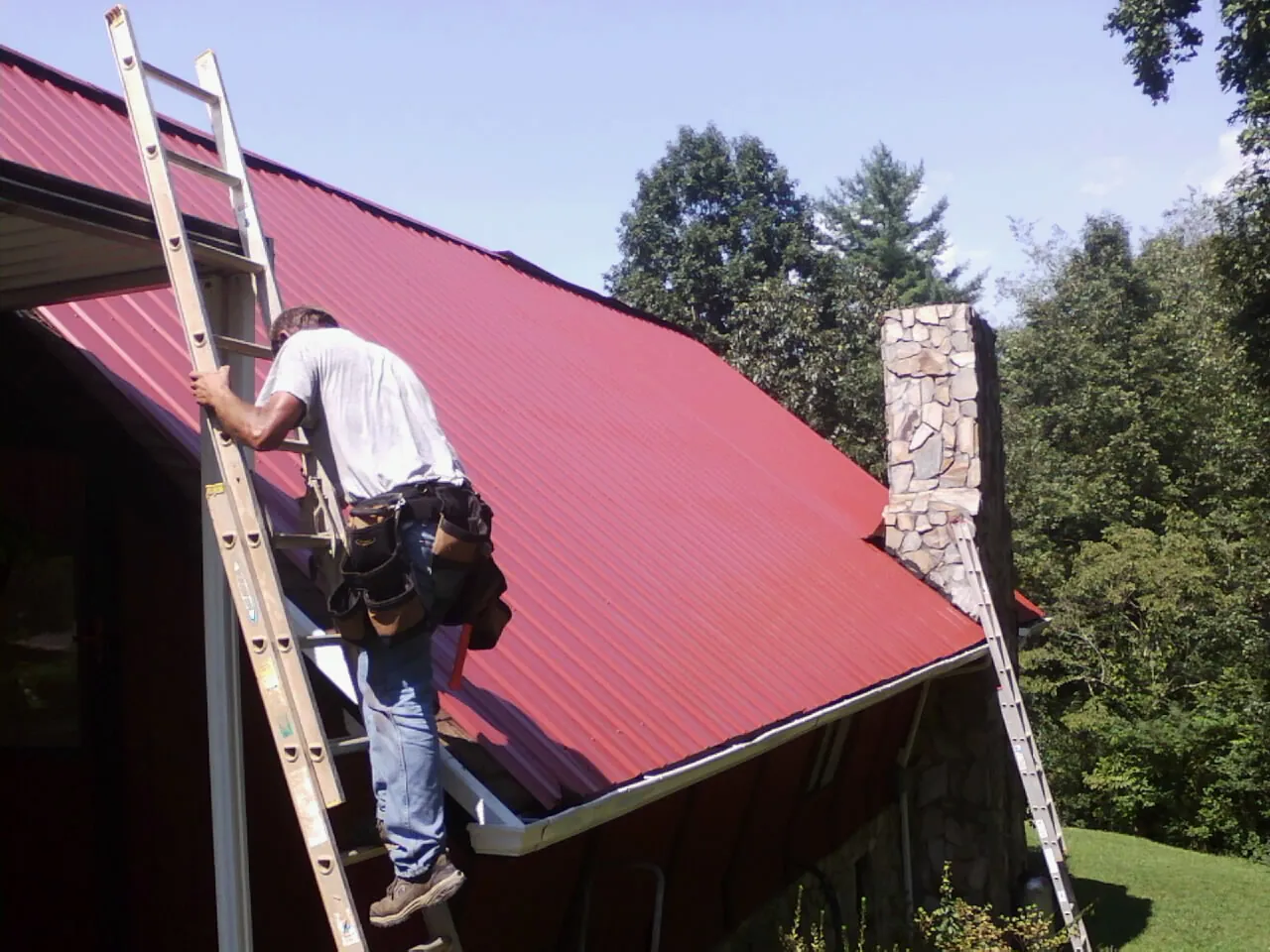 Roofer on a ladder beside a red metal roof and stone chimney — Soffit & Fascia Repair by Roofing Brothers in Townsend